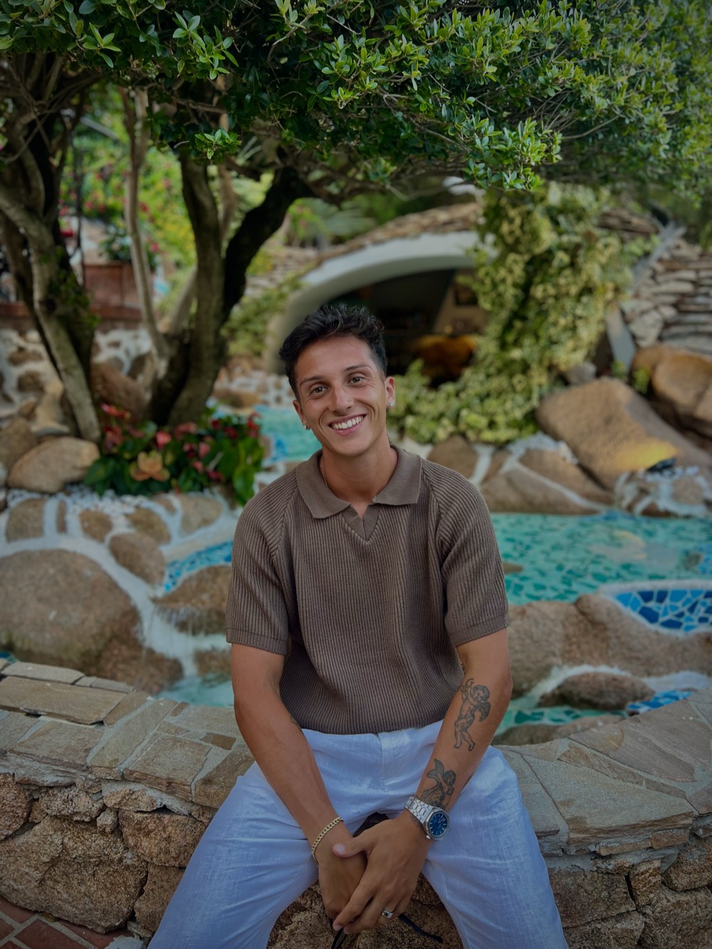Jacopo James Nardi smiling, sitting on a stone wall surrounded by greenery